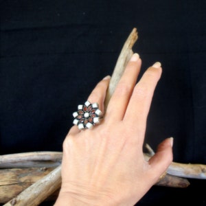 May include: A silver-toned ring with a floral design featuring white and reddish-brown stones. The ring is on a hand, with a dark background and a piece of driftwood.