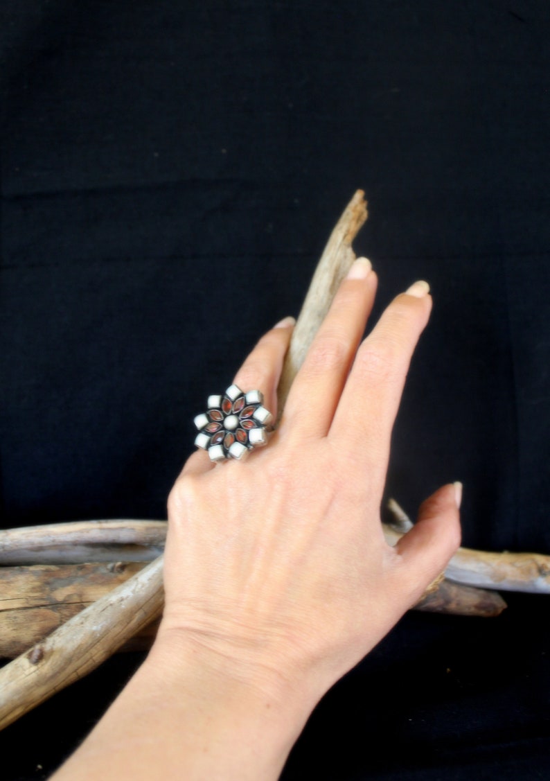May include: A silver-toned ring with a floral design featuring white and reddish-brown stones. The ring is on a hand, with a dark background and a piece of driftwood.