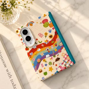 May include: A foldable phone case featuring a cream grid background with a colorful rainbow wave pattern. The design includes various small icons like flowers, stars, hearts, fruits, and animals. The case has a teal spine and a white camera cutout.
