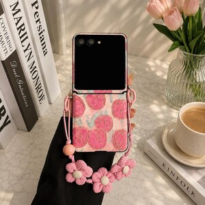 May include: A foldable smartphone case featuring a pink background with a repeating pattern of pink tulips. The case includes a matching pink strap adorned with three-dimensional fabric flowers and a single round bead for carrying.