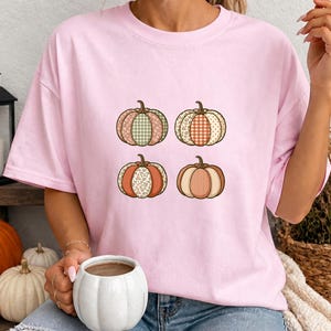May include: A light pink short-sleeve t-shirt featuring a graphic print of four pumpkins arranged in a square. Each pumpkin displays a different pattern, including green and white gingham, orange plaid, floral print, and vertical stripes in muted tones.