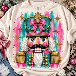 May include: A cream-colored crewneck t-shirt featuring a vibrant graphic of a nutcracker. The nutcracker wears a teal and pink hat with a pink bow, gold fringe epaulets, and has a black mustache and white beard, surrounded by gold starbursts.