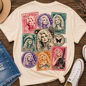 May include: A cream-coloured t-shirt featuring a collage of vintage-style postage stamps and circular graphics depicting Dolly Parton. The design includes text such as Dolly, 50c, USA, 1936, Coat of Many Colors, 1974, Jolene, and I Will Always Love You.