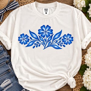 Op de afbeelding: Een wit T-shirt met korte mouwen en een centrale afbeelding van drie blauwe bloemen met gedetailleerde bloemblaadjes en bladeren. Het shirt heeft een ronde hals en een wit binnenlabel met de zwarte tekst Comfort Colors en een klein hartje.