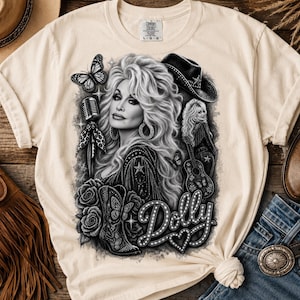 May include: A cream-coloured t-shirt featuring a black and white graphic print of a woman with voluminous hair, a cowboy hat, a microphone, a guitar, roses, a butterfly, and a cowboy boot. The text "Dolly" is printed in a marquee-style font.