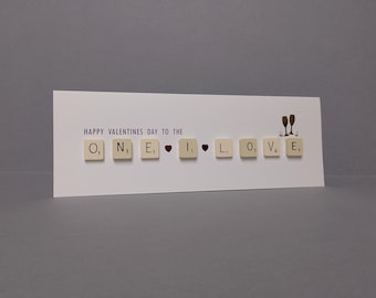 May include: A white Valentine's Day card with the message "HAPPY VALENTINES DAY TO THE ONE I LOVE" spelled out using Scrabble tiles. Two champagne glasses are depicted above the word "LOVE".