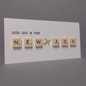 May include: A white card with the text "GOOD LUCK IN YOUR" and the word "NEW" spelled out with letter tiles. A small airplane tile is between the "W" and the "J" tile. The word "JOB" is spelled out with letter tiles.
