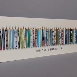 May include: A rectangular white greeting card featuring a row of forty vertical paper tubes arranged to resemble birthday candles. Each tube has a unique pattern and color, topped with a small gold flame. The text reads: HAPPY 40TH BIRTHDAY TIM.