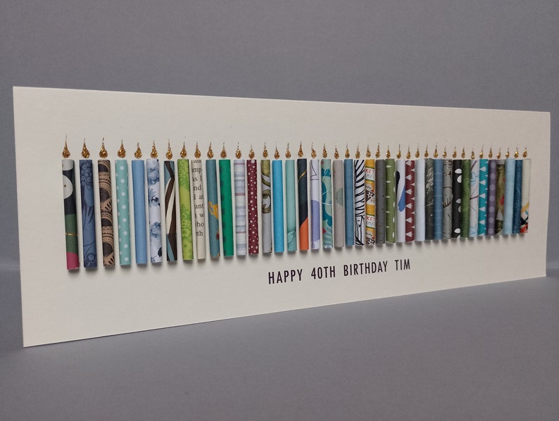 May include: A rectangular white greeting card featuring a row of forty vertical paper tubes arranged to resemble birthday candles. Each tube has a unique pattern and color, topped with a small gold flame. The text reads: HAPPY 40TH BIRTHDAY TIM.