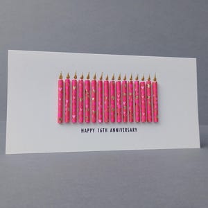 Puede incluir: Una tarjeta de felicitaci&oacute;n blanca con el texto "HAPPY 16TH ANNIVERSARY" presenta una fila de velas rosas con detalles dorados. Las velas tienen puntas doradas y un patr&oacute;n de coraz&oacute;n.