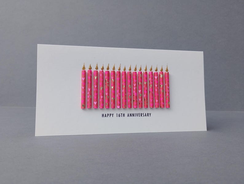 Puede incluir: Una tarjeta de felicitaci&oacute;n blanca con el texto "HAPPY 16TH ANNIVERSARY" presenta una fila de velas rosas con detalles dorados. Las velas tienen puntas doradas y un patr&oacute;n de coraz&oacute;n.
