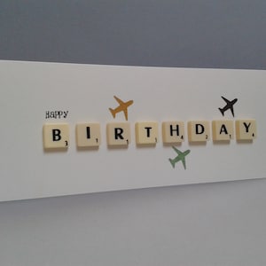 May include: A white card with the word "HAPPY" and the word "BIRTHDAY" spelled out using letter tiles. There are three paper airplanes on the card, one gold, one green, and one black.