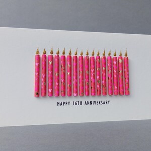 Puede incluir: Una tarjeta de felicitaci&oacute;n blanca con el texto "HAPPY 16TH ANNIVERSARY". La tarjeta presenta velas rosas decoradas con corazones dorados y llamas doradas brillantes.
