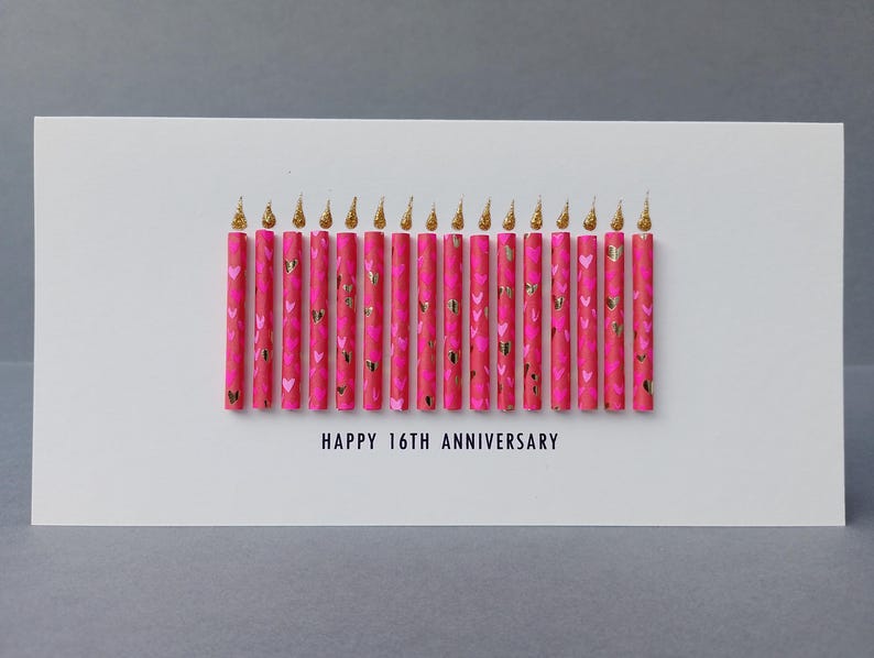 Puede incluir: Una tarjeta de felicitaci&oacute;n blanca con el texto "HAPPY 16TH ANNIVERSARY". La tarjeta presenta 16 velas rosas decoradas con corazones dorados y llamas doradas brillantes.