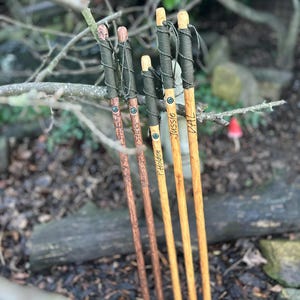 May include: Four wooden walking sticks with natural bark textures and varying wood tones. Each stick features a dark green cord wrapped around the top handle area, a small circular compass inlay, and custom text engraved vertically along the shaft.