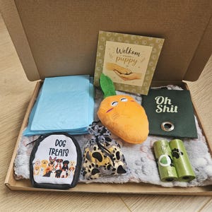 Op de afbeelding: Een puppy-starterset in een kartonnen doos met een lichtblauwe puppy-onderlegger, een pluche oranje wortelspeeltje, een groene poepzakjeshouder met de tekst Oh Shit, twee rollen poepzakjes, een beloningszakje met tekst en een Welkom puppy kaart.