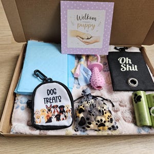 Op de afbeelding: Een puppy-cadeaubox met een lichtblauwe trainingsmat, een wit snoepzakje met hondenillustraties, een roze rubberen speeltje, een zwarte poepzakjeshouder met de tekst Oh Shit, een organza zakje met pootafdrukken en een Welkom puppy-kaart.