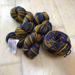May include: Four balls of yarn in shades of blue, gold, and brown. The yarn is arranged in a pile on a light wood surface. The label on one of the balls reads "Affairs Superwash Merino".