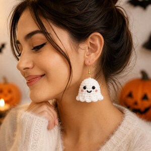 May include: A white crocheted ghost earring featuring a small, rounded body with a smiling face, black bead eyes, and pink stitched cheeks. The ghost is attached to a gold-toned metal hook finding, creating a dangling accessory for the ear.