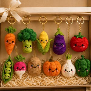 Op de afbeelding: Een set van elf gehaakte groente-sleutelhangers in een geschenkdoos. De collectie bevat een wortel, broccoli, maïs, aubergine, tomaat, erwtjes, radijs, aardappel, pompoen, knoflook en groene paprika, elk voorzien van een lachend gezichtje.