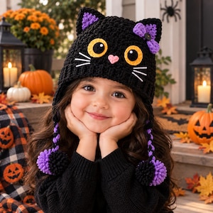 May include: A black crocheted cat hat featuring large yellow and black eyes, a small pink heart-shaped nose, white stitched whiskers, and purple inner ear details. The hat includes a purple bow and two long braided tassels with purple and black pom-poms.