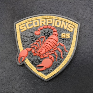 Könnte beinhalten: Ein schildförmiger Aufnäher mit einer erhabenen roten Skorpiongrafik auf schwarzem, strukturiertem Hintergrund. Der Rand ist gelb mit einem dekorativen Muster. Der Text SCORPIONS ist oben gelb aufgedruckt, mit den Buchstaben SS rechts.