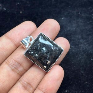 May include: A square pendant featuring a dark, textured stone with shimmering inclusions set in a polished silver-toned metal frame. The pendant includes a silver-toned bail at the top, designed for attachment to a necklace chain or cord.