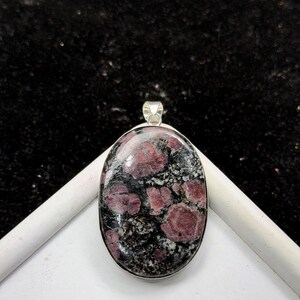 May include: An oval-shaped pendant featuring a polished gemstone with a mottled pattern of deep red, black, and white mineral inclusions. The stone is set in a smooth silver-toned metal bezel with a small integrated bail at the top for hanging.