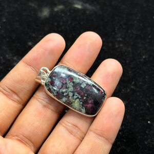 Puede incluir: Un colgante rectangular con una piedra preciosa pulida que presenta un patrón moteado de inclusiones de color rojo intenso, negro y verde claro, engastado en un marco de metal plateado con una pequeña anilla integrada en la parte superior.