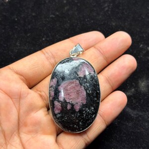 May include: An oval-shaped pendant featuring a polished black stone with prominent, irregular patches of deep pink and magenta mineral inclusions. The stone is set in a simple silver-toned metal frame with a small triangular bail at the top.