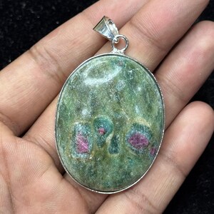 May include: An oval-shaped pendant featuring a polished green stone with embedded deep pink inclusions. The stone is set in a simple silver-toned metal frame with a matching silver-toned bail at the top for attachment to a chain or cord.