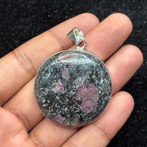 May include: A round pendant featuring a polished stone with a dark grey and black speckled matrix embedded with several distinct, irregular magenta-coloured mineral inclusions, set within a simple silver-toned metal frame and bail.