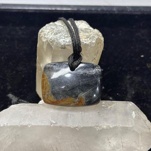 May include: A rectangular polished stone pendant featuring a dark gray and black surface with prominent streaks of golden-brown mineral inclusions, suspended from a simple black cord that loops through a hole at the top center of the stone.