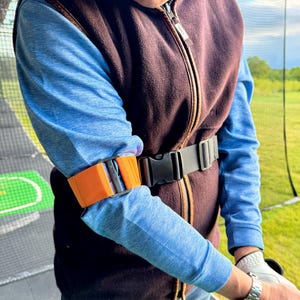 Peut inclure: Une sangle en nylon orange vif avec une boucle en plastique noir et des éléments de réglage en métal, fixée autour du bras sur un haut bleu à manches longues et un gilet polaire marron, avec une ceinture noire à boucle en plastique.