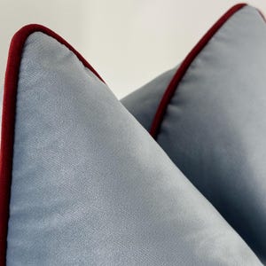 May include: Two light blue velvet throw pillows featuring a smooth, soft texture and a distinct deep red piping trim along the edges. The pillows are positioned at an angle, highlighting the contrast between the cool blue fabric and the dark red border.