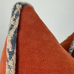 May include: A textured burnt orange pillow featuring a decorative edge trim. The trim consists of a woven pattern in shades of slate blue and cream, running vertically along the side of the pillow, contrasting with the solid orange fabric.
