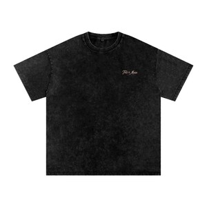 Puede incluir: Una camiseta negra de manga corta con acabado de lavado mineral. La prenda presenta un cuello redondo y el texto "FND BY MANZO" impreso en una fuente cursiva pequeña y de color claro en la parte superior izquierda del pecho.