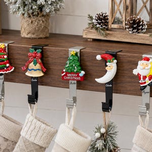 Può includere: Cinque ganci portacalza in metallo appesi a una mensola in legno, ognuno caratterizzato da un ornamento natalizio unico tra cui un albero di Natale, una campana dorata, un albero con la scritta Merry Christmas, un Babbo Natale e un sacco giallo.