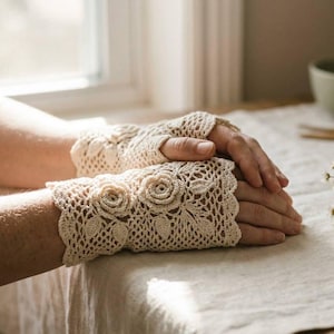 May include: A pair of cream-colored fingerless gloves featuring an intricate crochet lace pattern with three-dimensional floral motifs and scalloped edges. The gloves are crafted from fine yarn and cover the wrists and back of the hands.