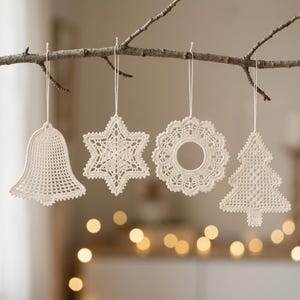 Op de afbeelding: Vier gebroken witte gehaakte ornamenten hangend aan een boomtak. De set bevat een bel, een zespuntige ster, een ronde krans met een opening in het midden en een dennenboomvorm. Elk stuk is gemaakt van fijn garen met gedetailleerde patronen.