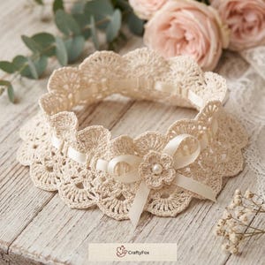 May include: A cream-colored crochet garter featuring a scalloped edge, a woven satin ribbon, and a central decorative bow with a small pearl accent. The item includes a small rectangular label with the text CraftyFox and a fox icon.