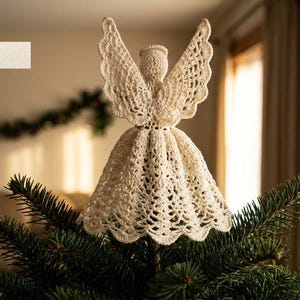 May include: A cream-colored crocheted angel tree topper featuring an intricate lace pattern on the skirt and wings, with a halo detail at the top. The figure is positioned atop a green pine tree branch. The text CraftyFox is visible.