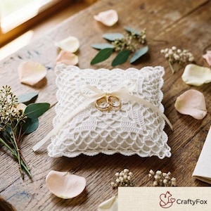 May include: A square white ring pillow featuring an intricate crochet lace overlay and a central cream-colored ribbon tied in a bow. Two gold wedding bands are secured by the ribbon. The pillow is surrounded by scattered pale pink rose petals.