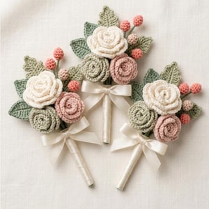 May include: Three handmade crochet buttonholes featuring cream, dusty pink, and sage green roses with matching leaves and small berries. Each floral arrangement is wrapped in white satin ribbon and finished with a tied white satin bow.