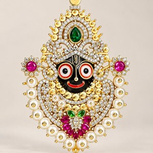 May include: A gold-toned pendant featuring a central black face with large white and red eyes and a red smile. The piece is adorned with clear crystals, white pearls, two pink stones on the sides, and green and pink stones at the bottom.