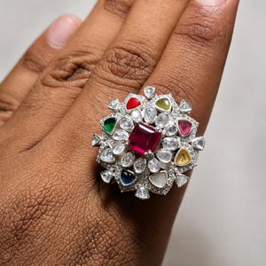 May include: A large silver ring featuring a central square-cut deep pink gemstone surrounded by a cluster of smaller, multi-colored stones including red, green, yellow, and blue gems, interspersed with numerous clear, irregular-shaped white stones.