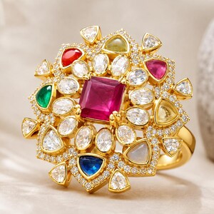 May include: A gold ring featuring a large square-cut red center stone surrounded by a cluster of smaller oval and triangular gemstones in red, green, blue, and clear white, all set within a frame of tiny pavé-set clear stones.