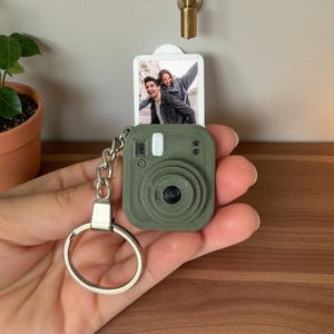 以下が含まれることがあります： 笑顔のカップルが写った引き出し式の長方形の写真インサートが付いた、オリーブグリーンのカメラ型キーホルダー。シルバーの金属製リングとチェーンが付いており、本体はヴィンテージのインスタントカメラを模した質感のあるプラスチック製です。