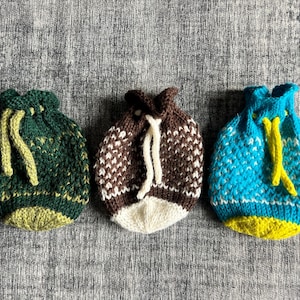 May include: Three knitted drawstring bags in shades of green, brown, and blue. The green bag has yellow drawstrings, the brown bag has white drawstrings, and the blue bag has yellow drawstrings. The bags are handmade and have a textured appearance.