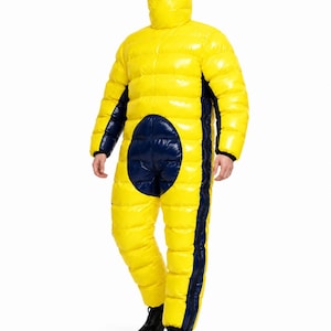 Op de afbeelding: Een felgeel, gewatteerd jumpsuit voor het hele lichaam met een glanzende afwerking, een capuchon met een horizontale oogopening, contrasterende donkerblauwe panelen aan de zijkanten en een grote cirkelvormige patch op de borst, gecombineerd met zwarte schoenen.